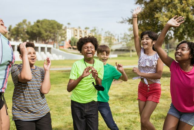Inclusive Summer Camp kids playing together in a sunny park, raising arms and laughing.